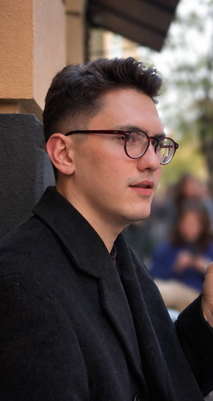 John Mitchell wearing glasses and a dark coat outdoors with blurred background