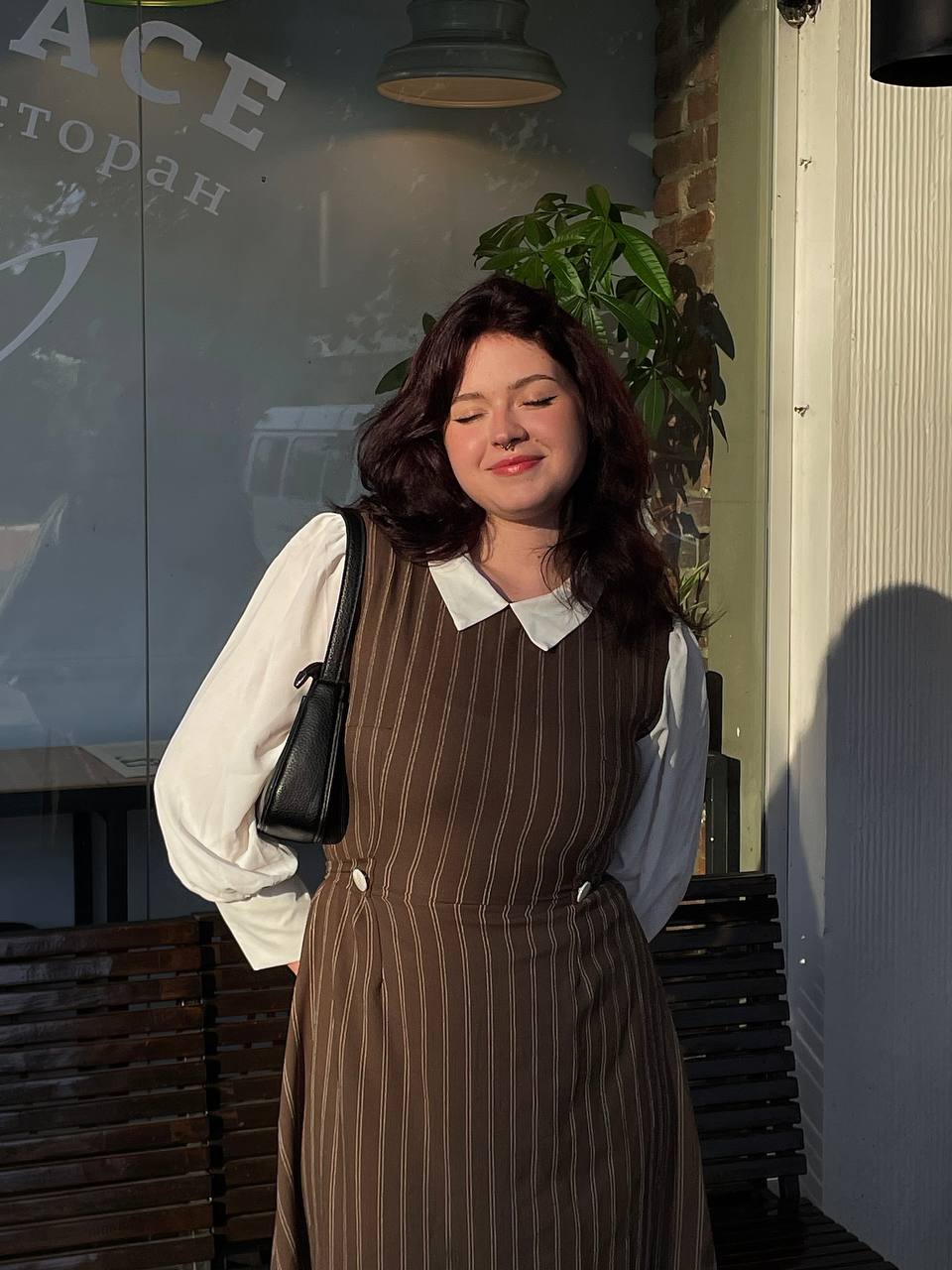 Emily Carter in a brown pinstripe dress standing outside a building with a plant in the background.