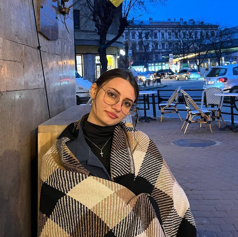 Karolina Sakun wearing glasses and a checkered coat sitting outdoors with a cityscape background